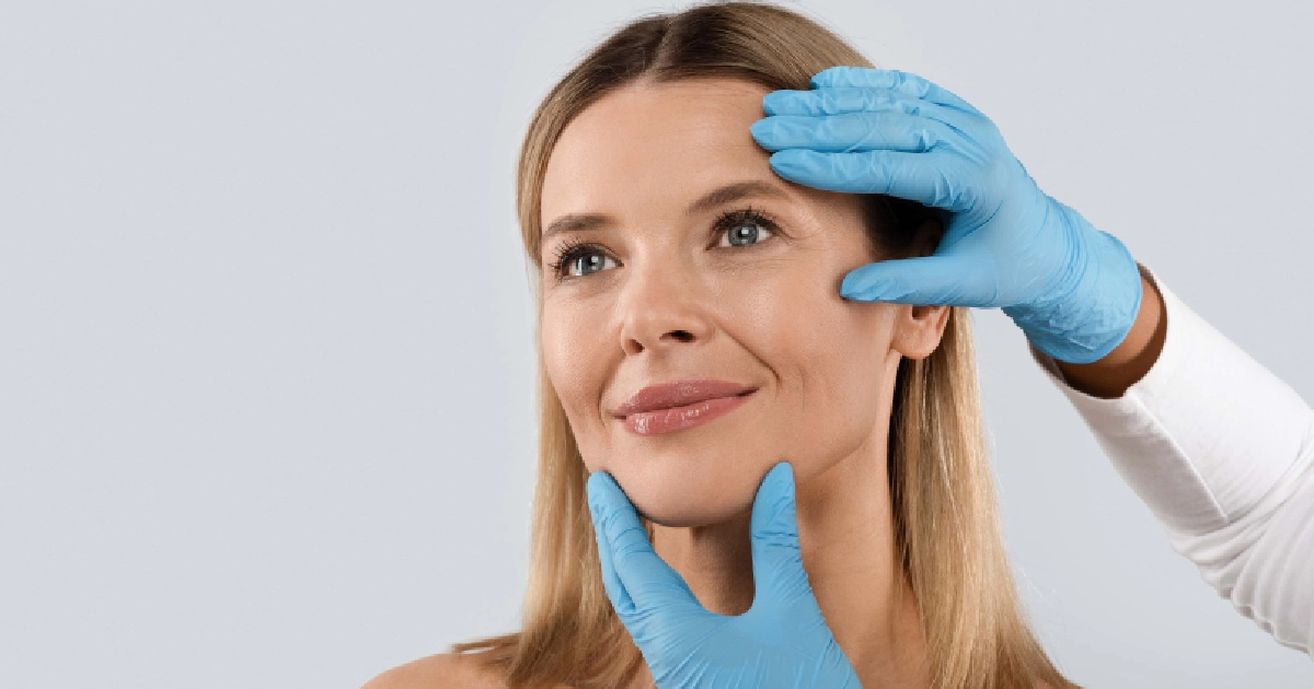 Smiling mature blonde woman with blue eyes being gently examined by a professional in blue gloves for Plastic Surgery in Whitefish, MT.-a--